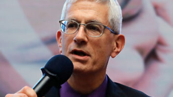 Andrew wears a dark suit jacket, purple shirt, and patterned neck tie. He is holding a microphone and talking.