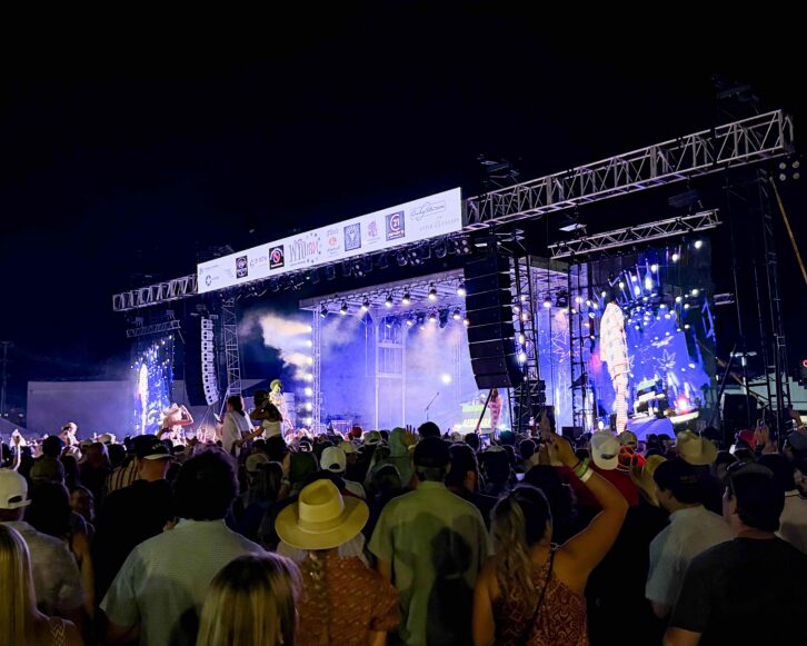 Large outdoor concert crowd facing a brightly lit festival stage equipped with suspended DAS Audio line array systems and large video screens.