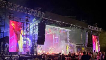 Outdoor live music stage at night with performers, large LED screens, colorful lighting, and DAS Audio line arrays positioned on both sides of the stage.