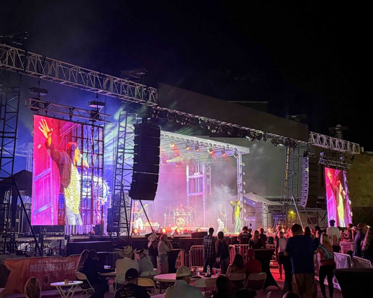 Outdoor live music stage at night with performers, large LED screens, colorful lighting, and DAS Audio line arrays positioned on both sides of the stage.