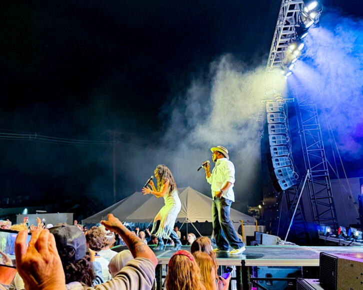 Two vocalists performing on an outdoor stage in front of a crowd, with a large DAS Audio line array suspended beside the stage.